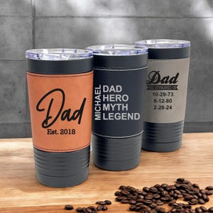 May include: Three stainless steel tumblers with different designs. The first tumbler has a brown leather wrap with the text "Dad Est. 2018". The second tumbler has a black leather wrap with the text "Michael Dad Hero Myth Legend". The third tumbler has a gray leather wrap with the text "Dad Was Established On 10-29-73 8-12-80 2-26-24".