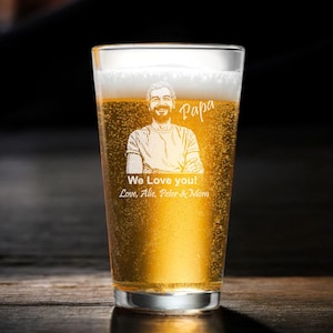 May include: A pint glass filled with beer, featuring an etched portrait of a man and the word "Papa". Below the portrait, the glass is inscribed with "We Love you!" and names. The beer has a foamy head.