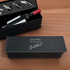 May include: Black leatherette wine box set with white stitching, containing wine tools and a bottle of wine. The box lid is engraved with the text "UNCLE MARC WILL YOU BE MY Godfather?"