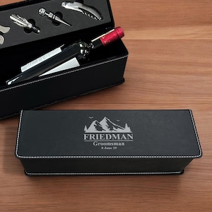 May include: Black leather gift box with a silver engraved mountain design and the text "FRIEDMAN  Groomsman 8 June 29". The box is open and a bottle of wine and wine accessories are inside.