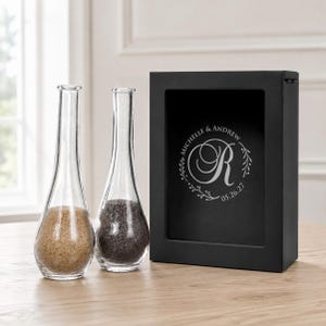 May include: Two clear glass bottles with narrow necks, one filled with tan sand and the other with dark gray sand, positioned next to a rectangular black box featuring a white engraved monogram with the names Michelle & Andrew and date 05.26.27.