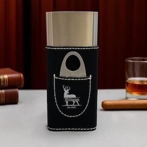 May include: A black leather cigar case with a silver-colored metal top and a built-in cigar cutter. The case has a pocket with a deer logo and the name "Williams" and the year "Est. 2025" stitched in white.