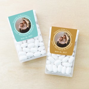 May include: Two clear plastic containers of mints with personalized wedding favor labels. The labels are round with a photo of a couple and the text "Thanks for coming! Love, Michelle & Andrew May 26, 2027". One label is teal and the other is gold.