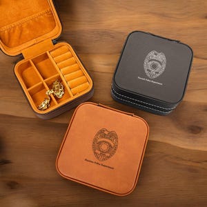 May include: Three square jewelry cases in brown, black, and orange leather. The open brown case reveals a yellow interior with ring slots and compartments. The black case has white stitching and a Newark Police Department badge. The orange case has a Houston Police Department badge.