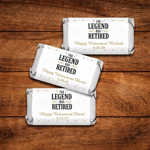 May include: Three wrapped chocolate bars with white labels and silver foil. The labels read "THE LEGEND HAS RETIRED" in black and gold, with personalized retirement messages and dates. The bars are arranged on a wooden surface.