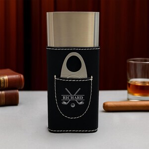 May include: A black leather cigar case with a silver-colored metal top. The case has a pocket with a golf-themed design and the name "RICHARD" engraved. A cigar cutter is in the pocket. A glass of amber liquid and a cigar are in the background.