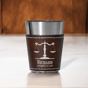 May include: A stainless steel shot glass with a brown leather sleeve. The sleeve features a white graphic of scales and the text "Richard, Attorney at Law". White stitching accents the leather.