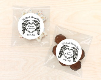 May include: Two clear plastic bags with white and brown chocolate candies. Each bag has a round sticker with a black and white illustration of two skeletal hands forming a heart shape. The stickers have the text "Til Death Do Us Part" and the names of the couples, "Monica & Patrick" and "Laura & David", along with their wedding dates.