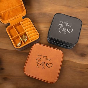 May include: Three small, square jewelry cases in brown, black, and orange leather. The black and orange cases have the text "love you DAD" with a stick figure drawing. The open brown case reveals an orange interior with ring slots.