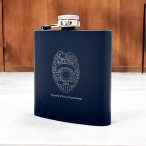 May include: A blue stainless steel flask with a silver cap and a silver engraved police badge with the text "HARTFORD" and "9346" and the words "Newark Police Department".