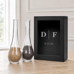May include: Two clear glass teardrop-shaped vases filled with sand, one containing tan sand and the other dark gray sand, positioned next to a rectangular black shadow box frame featuring the engraved letters D and F with the date 6.12.26.