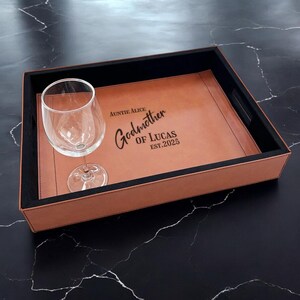 May include: A rectangular serving tray with a brown leather-like surface and black trim. The tray features the text "Auntie Alice Godmother of Lucas EST. 2025". A clear wine glass sits on the tray.