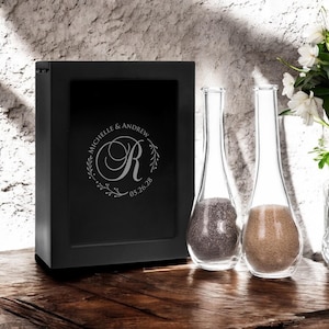 May include: Black wooden box with a silver engraved design featuring the names "Michelle & Andrew", the letter "R" and the date "05.26.28". Two clear glass vases with sand inside are in front of the box.
