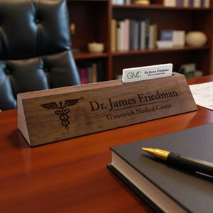 May include: A wooden desk nameplate with the name "Dr. James Friedman" and "Greenwich Medical Center" engraved. The nameplate also features a caduceus symbol and a slot for a business card. A pen and notebook are in the foreground.