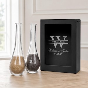 May include: Two clear glass vases with narrow necks, one filled with tan sand and the other with dark gray sand, positioned next to a rectangular black shadow box frame featuring the engraved text WILLIAMS, Dakota & John, and 04.26.27.