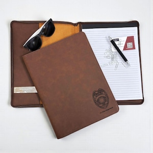 May include: A brown leather portfolio with a debossed police badge and the text "NY Police Department". The portfolio is open, revealing a notepad, pen, paper clips, and sunglasses.