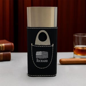 May include: Black leather cigar case with a stainless steel top and a pocket featuring an American flag design and the name "Richard". A cigar cutter is in the pocket. The case is on a table with a glass of amber liquid and a cigar.