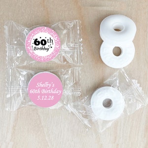 May include: Pink and white round favor bags with white mints and personalized labels. The labels say "60th Birthday" and "Shelby's 60th Birthday 5.12.28".