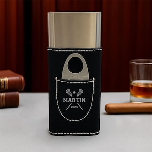 May include: A black leather cigar case with a silver-colored metal top and white stitching. A cigar cutter is in the front pocket. The pocket has the name "MARTIN" and the year "2025" printed on it.