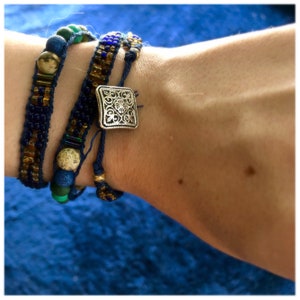 May include: Three beaded bracelets in shades of blue, gold, and green. The bracelets feature various bead sizes and textures, with a silver square clasp on one. The bracelets are designed to be worn together, creating a layered look.