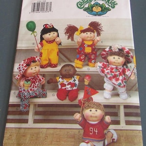 May include: A vintage Butterick sewing pattern featuring several Cabbage Patch Kids dolls. The dolls are dressed in various outfits, including yellow overalls, red and white patterned clothing, and a red jersey with the number 94. The pattern includes the text "ONE SIZE".