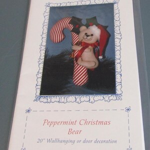 May include: A Christmas-themed wall hanging or door decoration featuring a teddy bear hugging a candy cane. The candy cane is red and white striped. The bear wears a Santa hat and a red ribbon. The text reads "Peppermint Christmas Bear, 20" Wallhanging or door decoration."