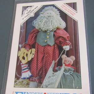 May include: A package featuring a 15-inch Country Yuletide Santa doll pattern. The Santa doll has a white beard, red plaid outfit, and a green vest. A small bear doll is also included in the pattern.