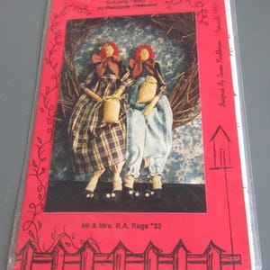 May include: A pattern packet featuring two rag dolls seated on a twig nest. The dolls have red hair, brown bows, and are dressed in plaid and denim outfits. The packet is in a red plastic sleeve with text that reads "SoLong Folks" and "Mr. & Mrs. R.A. Rags '32".