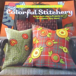 May include: Book cover featuring a red, orange, and green plaid pillow with colorful embroidered flowers. The book title is "Colorful Stitchery: 65 Hot Embroidery Projects to Personalize Your Home" by Kristin Nicholas.