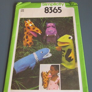 May include: Simplicity pattern 8365 featuring hand puppet designs. The puppets include a giraffe, hippo, dinosaur, and a bird. The giraffe puppet is orange with brown spots. The hippo is purple. The dinosaur is blue with a white zipper collar. The bird is green and yellow.