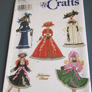 May include: A Simplicity Crafts sewing pattern featuring illustrations of dolls in elaborate, historical-style dresses. The pattern includes the text "9062" and "Julianna Blunt". The dolls are depicted in various colors, including red, black, and green.