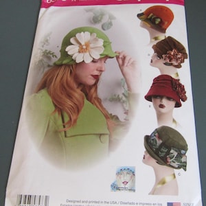 May include: A Simplicity sewing pattern envelope, number 8573, featuring illustrations of five different cloche-style hats. The main image shows a green hat with a large white flower, while smaller images display variations in orange, brown, red, and floral fabric.