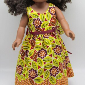 May include: A doll wearing a yellow dress with a leaf and geometric pattern, a matching headband, and a burgundy ribbon belt. The doll has dark curly hair and is set against a white background.