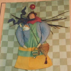 May include: A handmade fabric doll with a whimsical design. The doll wears a green dress, yellow skirt, and blue jacket. It has a red collar, a flower on its head, and a small bag that says "SEED". The doll is mounted on a green stand.