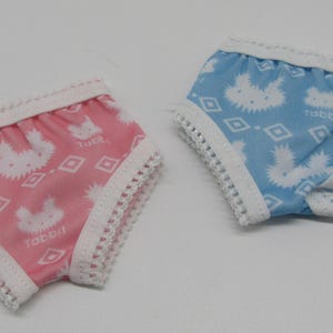May include: Two pairs of doll underwear. One pair is pink with white rabbit and diamond patterns, and the word "Tabbii". The other pair is blue with white rabbit and diamond patterns, and the word "Tabbii". Both have white lace trim.