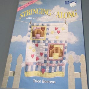 May include: A book cover titled "Stringing Along with Vanessa + Ann" features a quilt design with cat and heart appliqués. The quilt is displayed against a sky background with a white picket fence. The designer's name, Trice Boerens, is also visible.