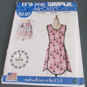 May include: A McCall's sewing pattern envelope titled It's Sew Simple, style L9417, size S-L. The front displays a pink apron with a black Eiffel Tower print and black trim, alongside a tiered skirt illustration. Text includes 2 Yards and Made in USA.
