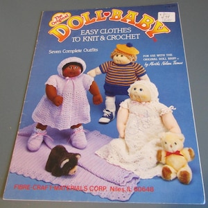 May include: A vintage book cover titled "The Original Doll Baby" with the subtitle "Easy Clothes to Knit & Crochet." The cover features three dolls in handmade outfits, including a crocheted dress and a striped shirt. A small teddy bear is also present.
