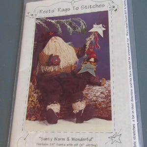 May include: A clear plastic bag containing a craft pattern. The pattern features a Santa Claus doll with a long white beard, holding a star-shaped ornament. The text reads "Santy Warm & Wonderful" and "Reets' Rags To Stitches".