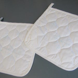 May include: Two square white quilted pot holders featuring a subtle light blue embroidered snowflake pattern. Each pot holder has a matching white fabric border and a small fabric loop attached to one corner for convenient hanging storage.