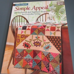 May include: A softcover book titled Simple Appeal: 14 Patchwork and Appliqué Projects for Everyday Living by Kim Diehl. The cover features a colorful quilted table runner with floral appliqué and geometric patterns draped over a wooden table.