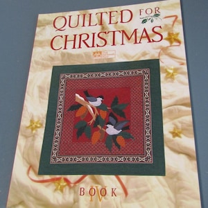 May include: A softcover book titled Quilted for Christmas Book IV. The cover features a cream-colored quilted background with gold stars and a central square image of two birds perched on a branch with pinecones, set against a red and green border.
