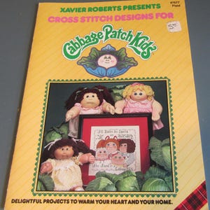 May include: A yellow book cover with the text "Xavier Roberts Presents Cross Stitch Designs for Cabbage Patch Kids." The cover features Cabbage Patch Kids dolls and a cross-stitch design. The text "Delightful Projects to Warm Your Heart and Your Home" is at the bottom.