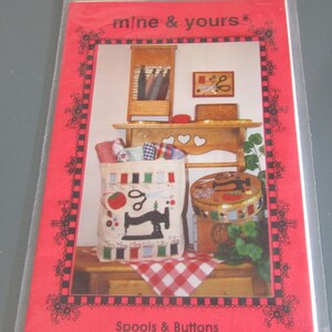 May include: A craft pattern packet titled mine & yours Spools & Buttons in a clear plastic sleeve. The cover features a photograph of a fabric bag with a sewing machine graphic, a round tin, and wooden shelves decorated with sewing supplies.