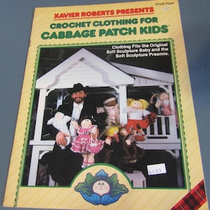 May include: A book cover with the title "Xavier Roberts Presents Crochet Clothing for Cabbage Patch Kids." The cover features a photo of a man holding several Cabbage Patch Kids dolls in front of a white structure. The cover has a yellow and green border.