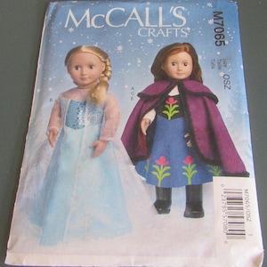 May include: A McCall's Crafts sewing pattern featuring two dolls dressed in costumes. One doll wears a light blue dress with a snowflake design, and the other doll is in a blue dress with a purple cape. The pattern cover has the text "McCall's Crafts" and "M7065".