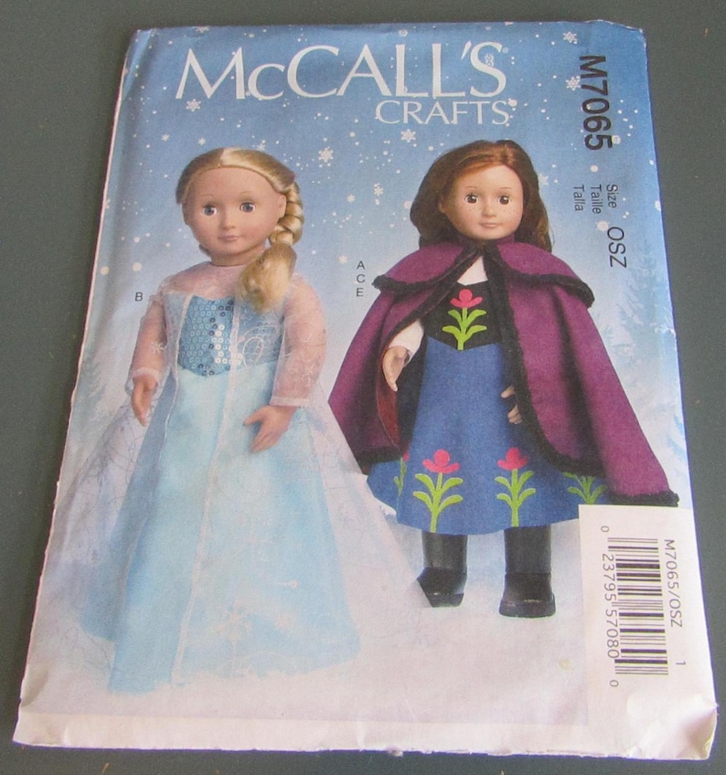 Puede incluir: Un patr&oacute;n de costura McCall's Crafts con dos mu&ntilde;ecas vestidas con disfraces. Una mu&ntilde;eca lleva un vestido azul claro con un dise&ntilde;o de copo de nieve, y la otra mu&ntilde;eca lleva un vestido azul con una capa morada. El patr&oacute;n tiene el texto "McCall's Crafts" y "M7065".
