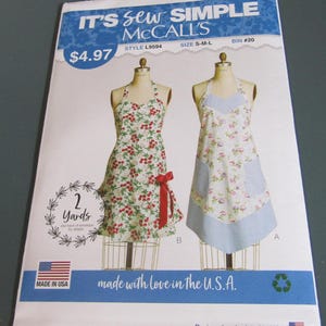 May include: A McCall's sewing pattern envelope titled IT'S SEW SIMPLE featuring two apron designs on mannequins. One apron has a red cherry print with a red bow, and the other has a floral pattern with blue gingham accents. Text includes 2 Yards.