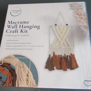 May include: Macrame wall hanging craft kit. The box features a finished macrame wall hanging with cream and brown tassels. The kit includes easy DIY assembly instructions. Finished size is approximately 7.8 x 17.7 inches.