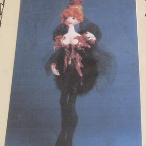 May include: A handmade doll with red hair, wearing a black dress with a feathered skirt and a red hat. The doll has a white chest and is posed against a blue background.